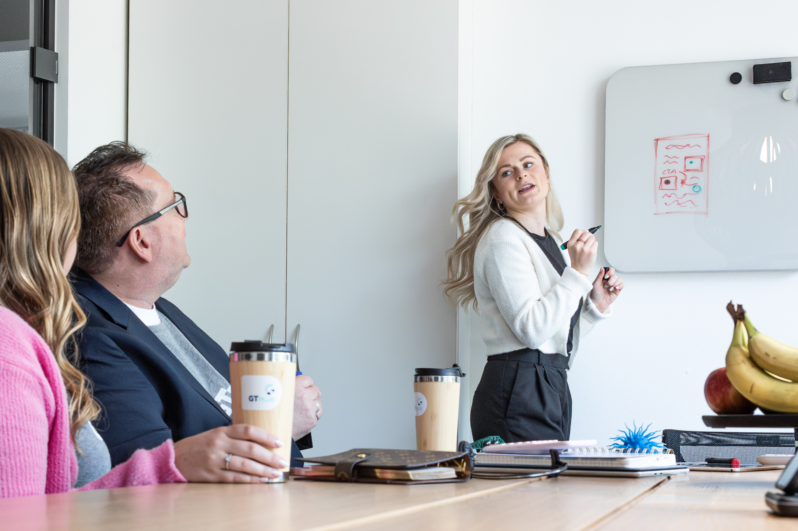 Frau hält einen Vortrag und erklärt zwei weiteren Personen etwas. Sie hält einen Stift in der Hand, das Whiteboard ist hinter ihr.