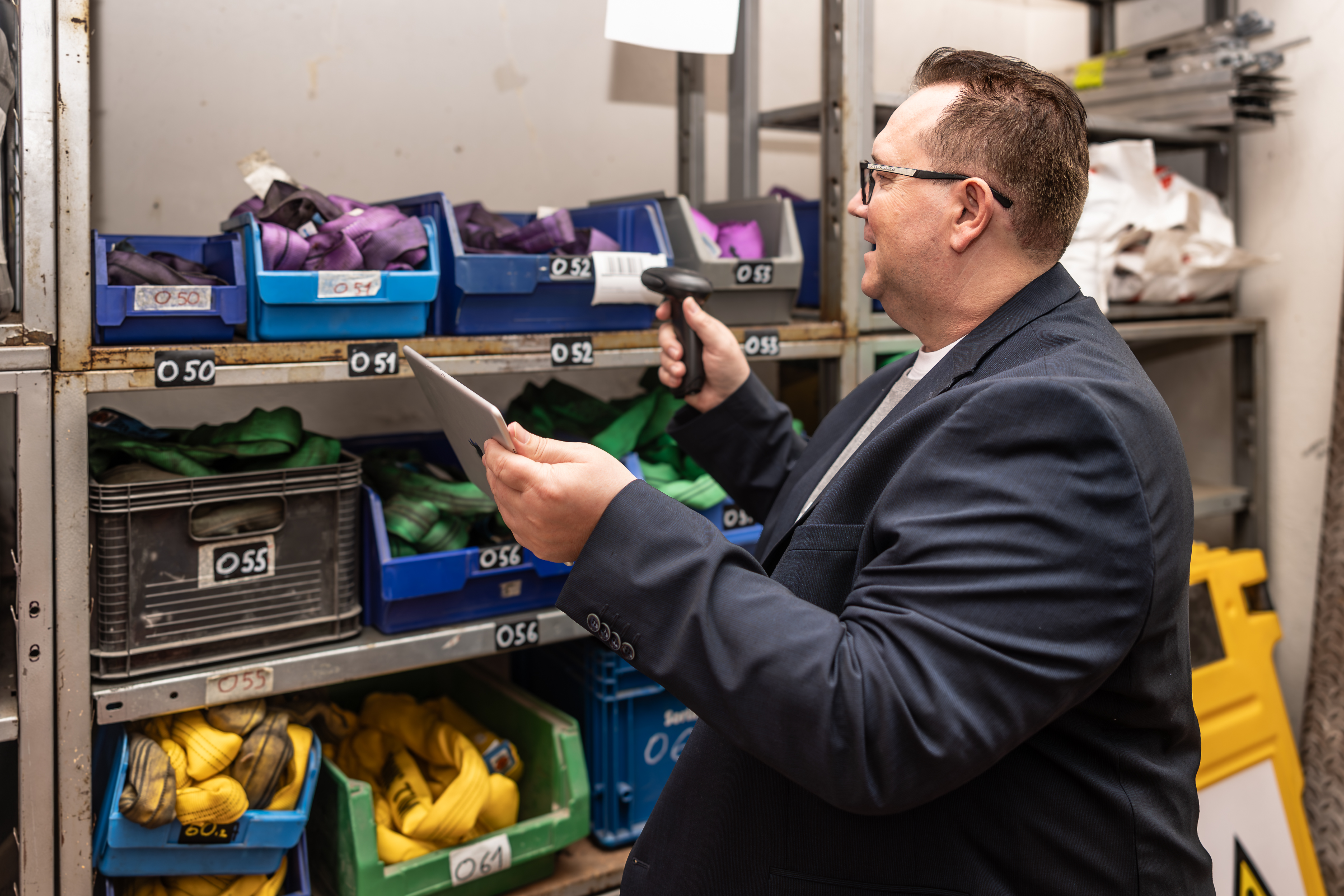 Mann arbeitet mit einem Scanner im Lager und sannt Barcodes ab. Er hält zudem ein Tablet in der Hand und überprüft den Lagerbestand.