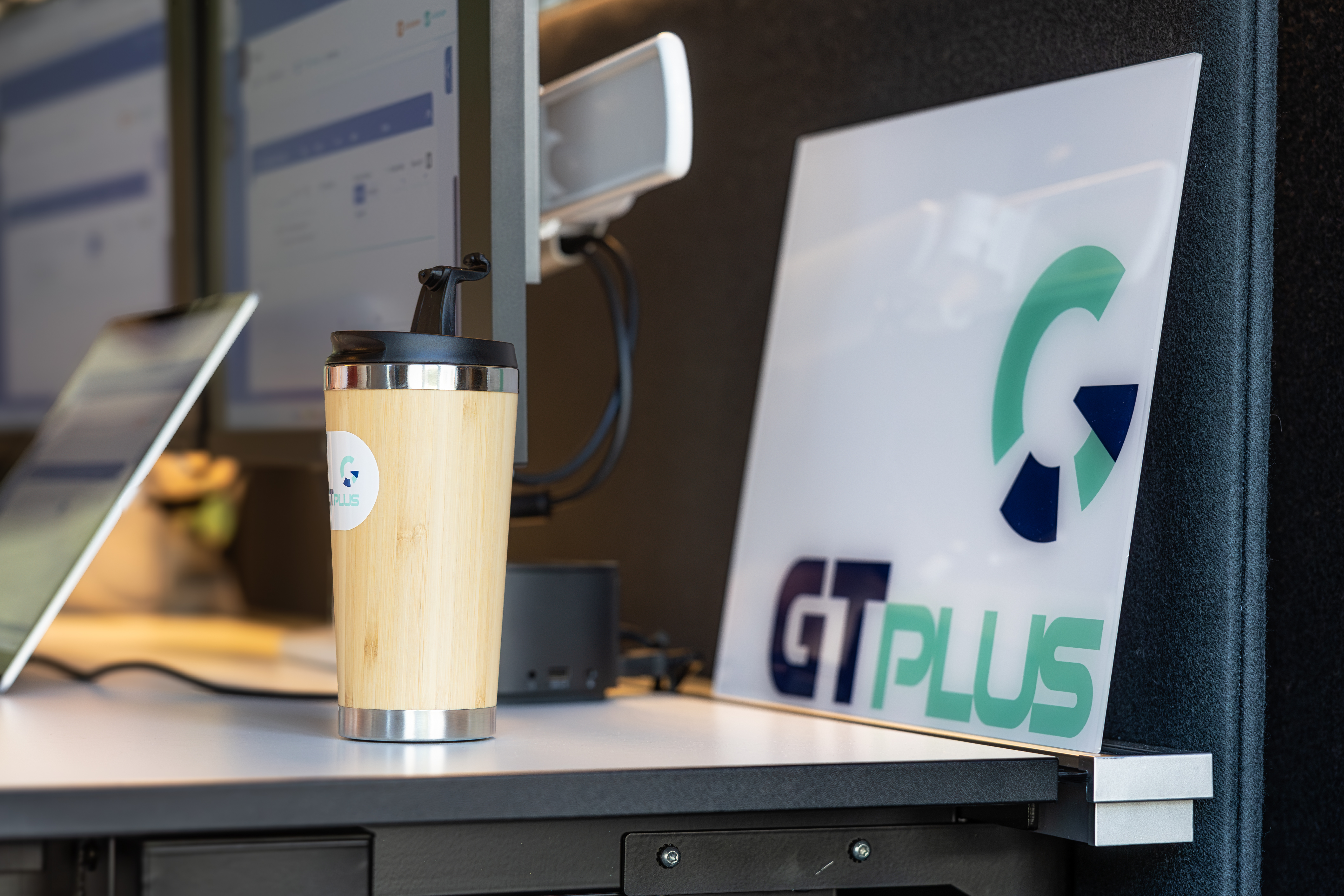 Arbeitsplatz mit einem Schreibtisch, Kaffeebecher, Laptop und Firmenlogo Schild von GT-Plus.