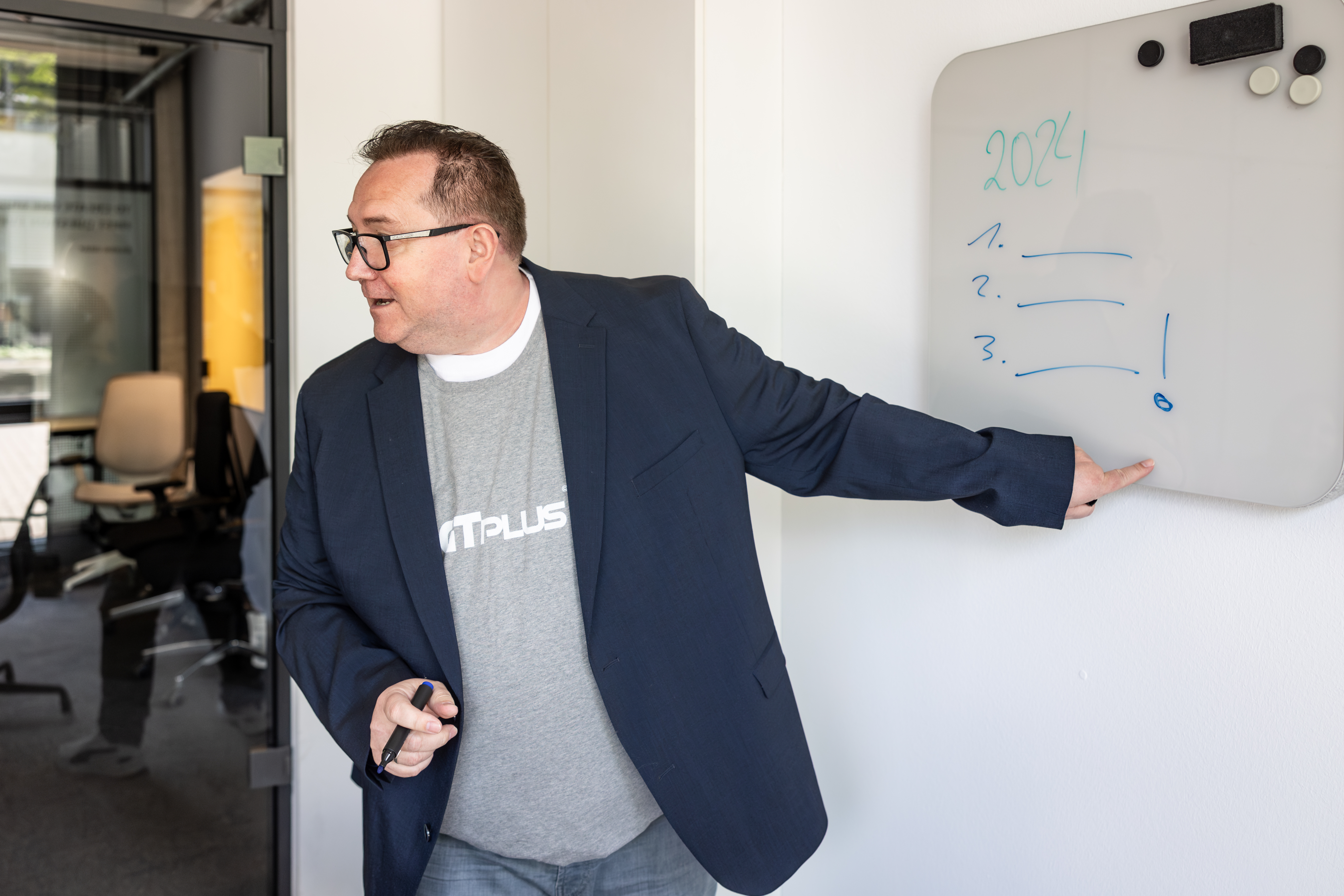 Mann hält einen Vortrag und zeigt mit dem Finger auf ein Whiteboard mit einer Schrift. Er schaut seine Zuschauer (welche nicht mit im Bild sind) an und spricht.