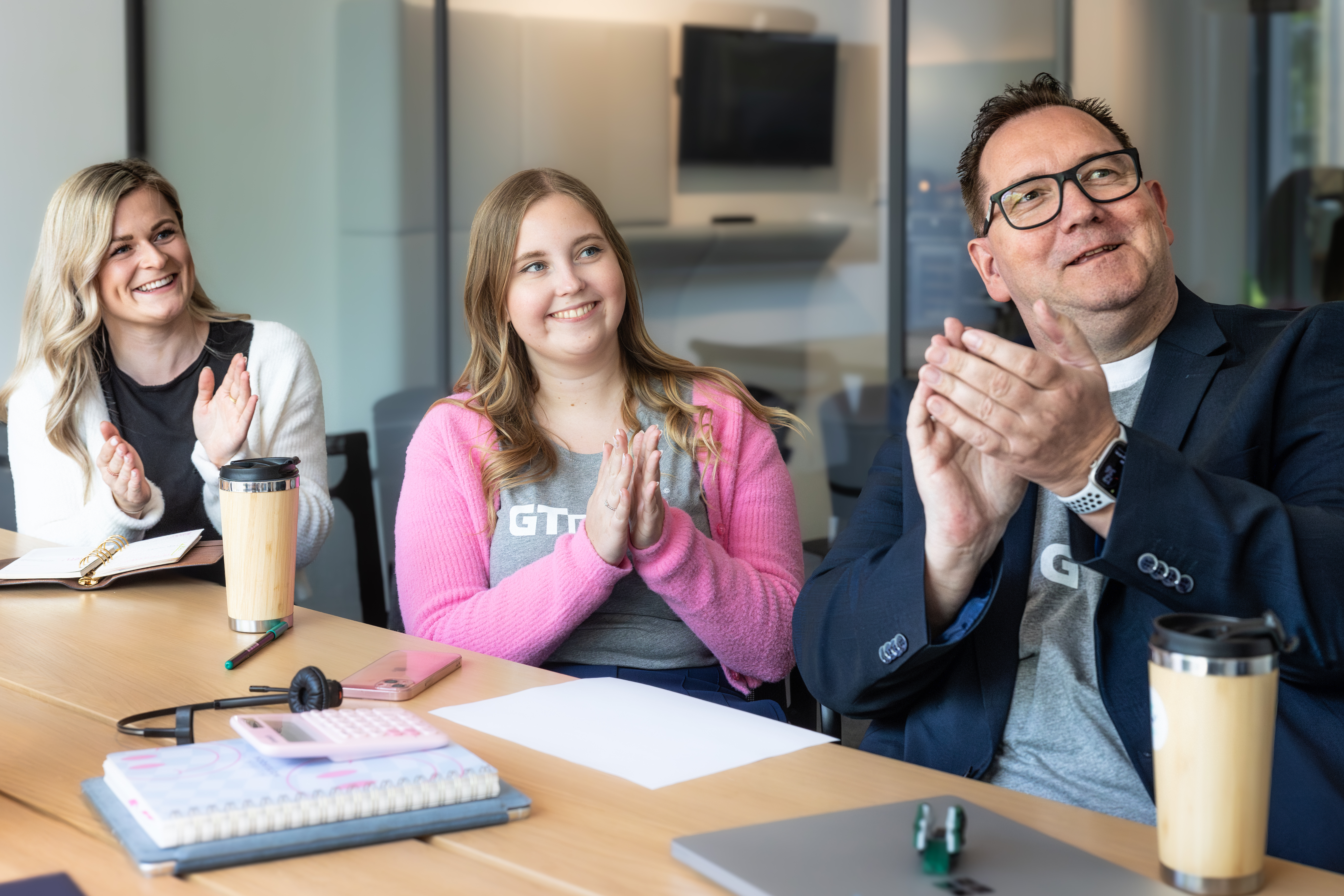 Drei Personen sitzen in einem Besprechungsraum und klatschen, lächeln und schauen alle in eine Richtung.