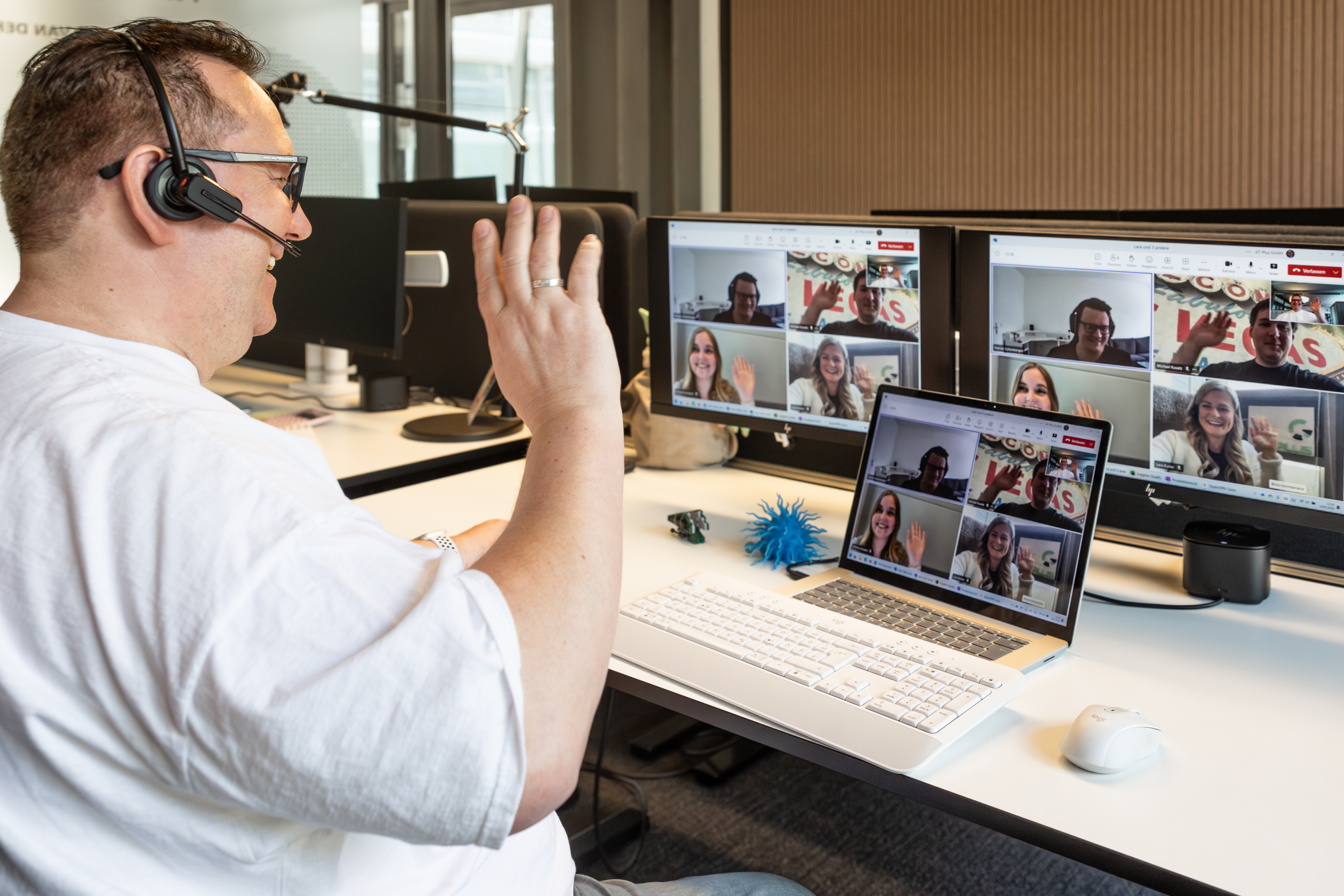 Mann sitzt an einem Schreibtisch mit Computern und ist gerade in einem TEAMS-Meeting mit vier weiteren Personen. Alle Personen winken und lächeln sich an.