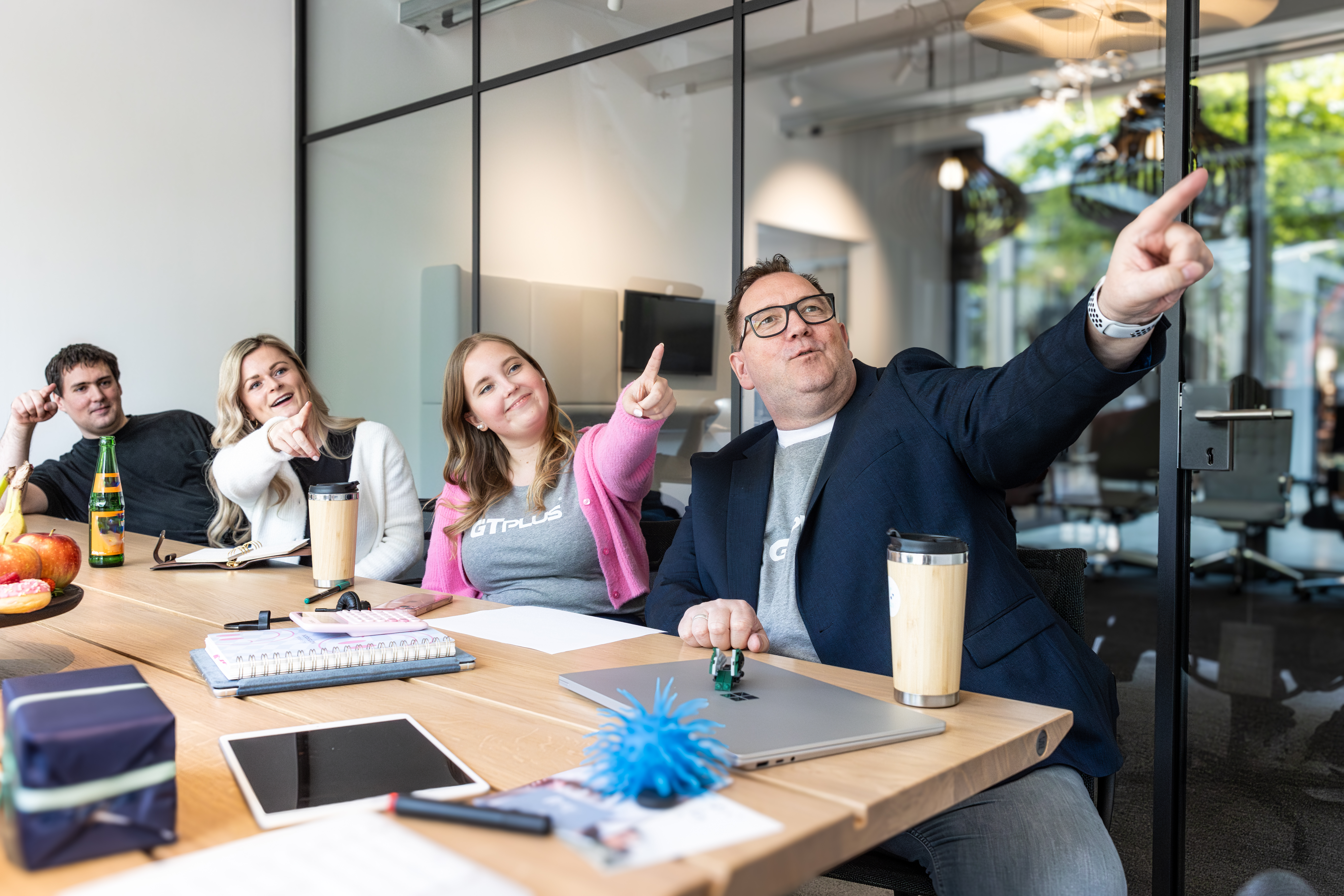 Vier Personen sitzen in einem Besprechungsraum am Tisch und zeigen alle mit dem Zeigefinger in eine Richtung, alle sind fröhlich.